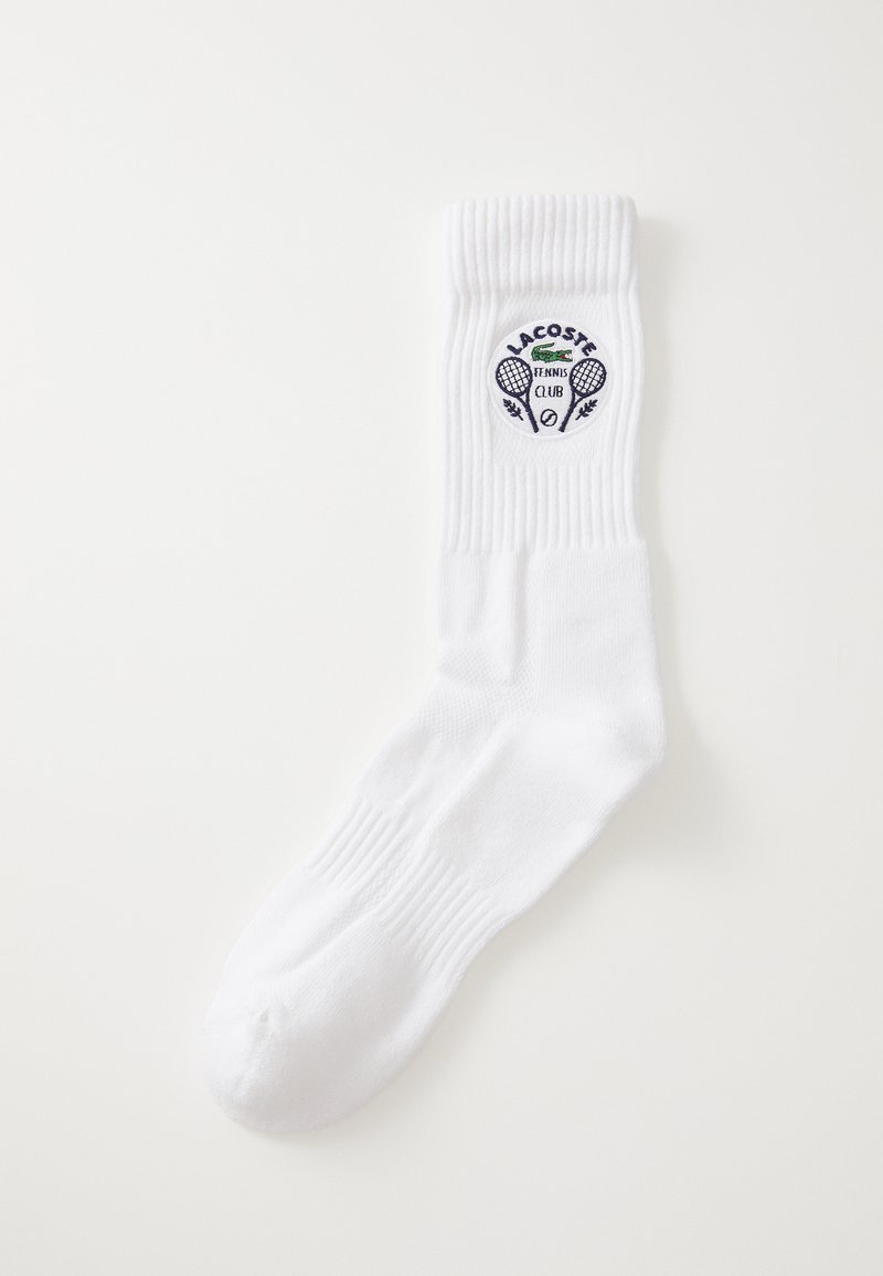 Bílá bavlněná ponožka s žebrovaným lemem, na které je kulaté logo Lacoste Tennis Club v námořnické modři, včetně zkřížených raket a vavřínových akcentů.