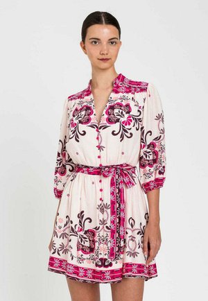 Donna che indossa un abito bianco con motivi floreali rosa e neri, maniche a tre quarti, davanti con bottoni e cintura coordinata in vita su sfondo uniforme.