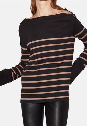 Vrouw draagt een zwart langärmlig shirt met horizontale beige strepen en leren broek, ritssluitingdetail op de schouder, handen in de zij.