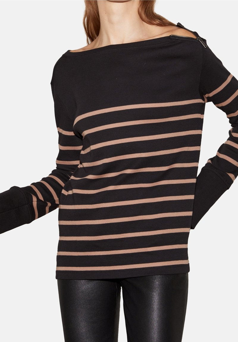 Femme portant un haut noir à manches longues avec des rayures beiges horizontales et un pantalon en cuir, détail de fermeture éclair sur l'épaule, mains sur les hanches.