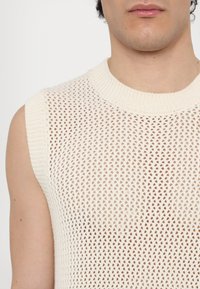 Homme portant un haut en maille crème sans manches avec col côtelé, visage partiel et torse supérieur visibles sur un fond blanc uni.