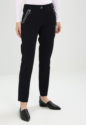 Kvinde iført slanke sorte bukser med sølvfarvede lynlåslommer foran og sorte loafers med spids tå, stående mod en hvid baggrund.