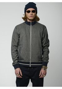 Giacca grigia testurizzata con zip frontale, dettagli blu navy, polsini a costine e due tasche laterali. Indossata con un berretto blu navy e occhiali da sole.