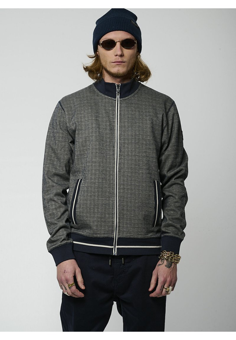 Giacca grigia testurizzata con zip frontale, dettagli blu navy, polsini a costine e due tasche laterali. Indossata con un berretto blu navy e occhiali da sole.