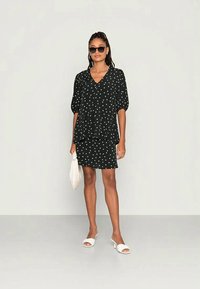 Blusa preta com bolinhas brancas e mangas bufantes, sobre uma saia correspondente. A modelo usa sandálias brancas e segura uma bolsa branca.