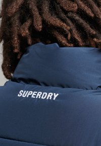 Giacca isolante blu navy con colletto alto, caratterizzata da un logo ricamato bianco "SUPERDRY" sul retro e una texture di tessuto liscia e lucida.