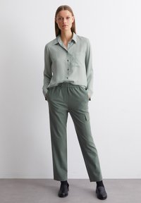 Light green button-up shirt with a chest pocket, paired with dark green elastic-waist pants featuring a side pocket. Black shoes.