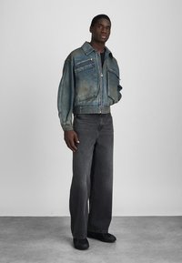 Mees seisab seljas pleekinud sinine lukuga teksajakk koos plaastrikotidega, laiad mustad teksad ja mustad kingad, ühtlasel hallil taustal.