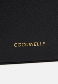 Černá texturovaná kůže s reliéfní strukturou zrna, uprostřed zlaté logo "COCCINELLE". Žádné další kovové doplňky nebo akcenty nejsou viditelné.