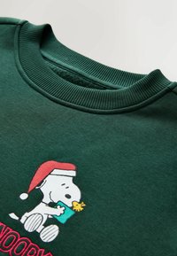 Zöld pulóver bordás gallérral, amelyen egy grafika látható Snoopy-ról, amint Mikulás sapkát visel, és egy ajándékot tart a kezében. Alatta rózsaszínű "SNOOPY" felirat található.