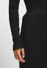 Czarna, teksturowana sukienka z długimi, dopasowanymi rękawami. Materiał posiada falbaniasty wzór wzdłuż rękawów i ciała, z wcięciem w talii na pasku.