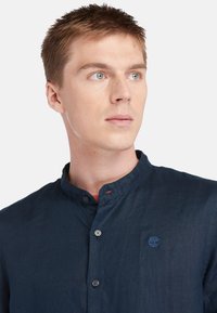 Camicia blu navy con colletto alla cinese, texture in tessuto, due bottoni visibili e un piccolo logo ricamato sul petto.