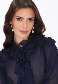 Tengerészkék áttetsző blúz fodros részletekkel, magas nyakkal és megkötőkkel, valamint hosszú ujjakkal. Nagy gyöngy fülbevalókkal díszítve.