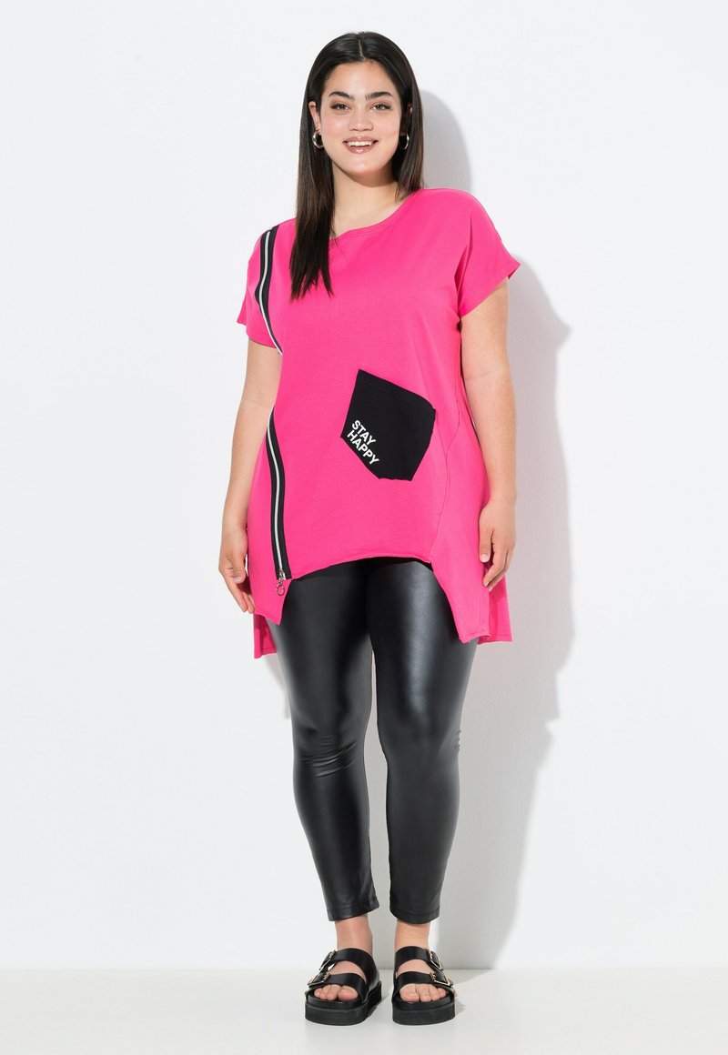 Roze tuniek met korte mouwen, zwarte zak met de tekst "STAY HAPPY", zijritsdetail, gecombineerd met zwarte faux leren leggings.