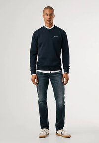 Sweatshirt de algodão azul-marinho com decote redondo, punhos e bainha canelados, usada por cima de uma camisa branca com gola, combinada com jeans escuros e ténis.