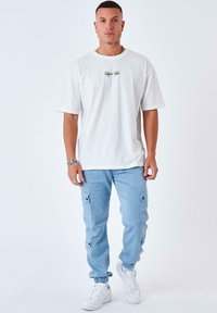 Camiseta blanca de gran tamaño con logo negro, pantalones cargo azul claro con bolsillos y zapatillas blancas; tejido suave y ajuste relajado.