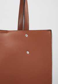 Poignée de sac fourre-tout en cuir marron texturé, fixée avec deux rivets argentés, présentée sur un fond neutre.