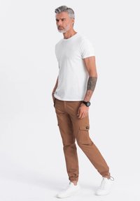 Bruine cargo broeken met elastische boorden, zijzakken en een zwart logo patch, gecombineerd met een aansluitend wit T-shirt en witte sneakers.