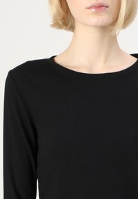 Crna rebrasta majica dugih rukava s okruglim izrezom, s glatkom teksturom i prilagođenim dizajnom, ističe dekolte i područje ramena.