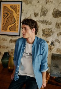 Camisa de denim azul claro sobre una camiseta blanca con impresión negra, combinada con vaqueros oscuros. Fondo de madera y piedra con elementos decorativos.