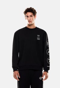 Černá mikina s kulatým výstřihem, s bílými grafickými doplňky na hrudi a levém rukávu. Vyrobena z měkké, texturované látky.