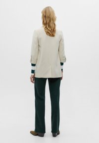 Blazer beige con maniche arrotolate sopra pantaloni verde a gamba larga. Stivaletti alla caviglia marrone scuro. Tessuto liscio con design minimale e dettagli di sartoria discreti.