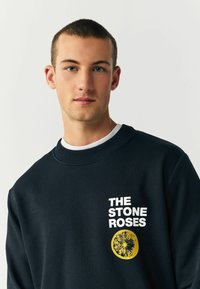 Mørkeblå sweatshirt med rund hals. Har hvid tekst, der står "THE STONE ROSES", samt en gul grafik af en blomst. Blød tekstur.