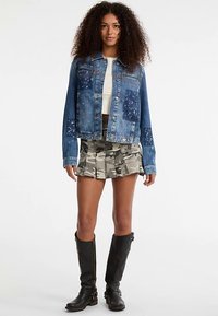 Veste en jean à lavage bleu, texture usée ; associée à une jupe plissée camouflage et des bottes noires montant jusqu'aux genoux.