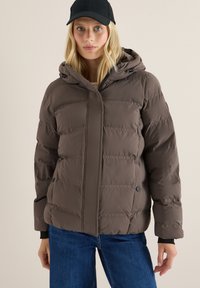 Veste matelassée marron avec une capuche, présentant une fermeture éclair à l'avant et des sections matelassées horizontales. Portée avec un jean bleu et complétée par une casquette noire.