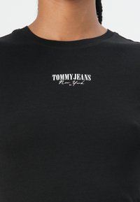 Melnas kokvilnas T-krekls ar apaļu kakla izgriezumu. Priekšpusē ir balts "TOMMYJEANS New York" logs, kas izdrukāts uz krekl. Gluda tekstūra.