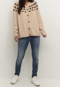 Ljusrosa stickad cardigan med bruna geometriska mönster på axlarna, utrustad med stora knappar och ribbade ärmar, i en kombination med blå jeans och vita sneakers.