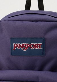 Gros plan sur un sac à dos JanSport violet montrant la texture du tissu et le patch du logo de la marque cousu sur la poche avant.