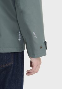 Veste verte imperméable avec une texture lisse, comportant un logo GORE-TEX, des accents argentés et des détails de poignet à boutons.