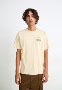 Beige cotton t-shirt with short sleeves and a round neckline. Features a small green graphic on the left chest. Smooth texture, relaxed fit.