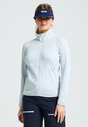 Una mujer luce una chaqueta blanca con cremallera, unos pantalones azul marino y una visera SLAM azul marino sobre un fondo liso y claro.
