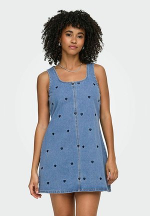 Görög hajú nő ujjatlan farmerruhában, amelyen apró fekete szívminták vannak, egy sima világos háttér előtt állva.