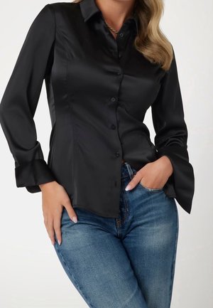 Vrouw draagt een nauwsluitende zwarte satijnen blouse met knopen en lange mouwen, gecombineerd met een blauwe spijkerbroek, met één hand in de zak en de andere rustend op de heup.