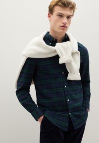 Groen en marineblauw geruit overhemd met knopen en lange mouwen, gecombineerd met een crèmekleurige gebreide trui die over de schouders hangt.