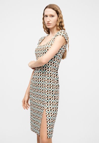 Γυναίκα με μακριά κυματιστά μαλλιά, φορώντας εφαρμοστό φόρεμα με γεωμετρικό μοτίβο, κοντά μανίκια και ψηλό άνοιγμα στο πλάι, στέκεται μπροστά σε ένα απλό φόντο.