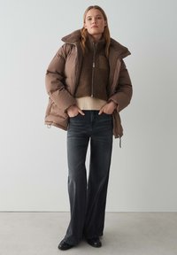 Doudoune marron avec un col en fourrure sur un haut en polaire marron. Jeans gris évasés et bottines noires. Style simple et décontracté.