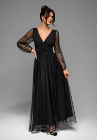 Abito maxi nero con scollatura a V profonda, maniche lunghe trasparenti e vita aderente, caratterizzato da una gonna fluente e un sovrapposto in tulle a strati.