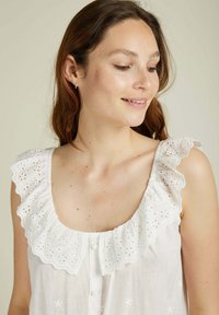 Blouse blanche sans manches avec un volant en coton brodé à bord festonné et des détails de boutons, mettant en avant des motifs floraux sur le tissu.