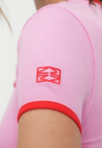 Ροζ εφαρμοστό τοπ με ριμπ ύφανση και κόκκινες λεπτομέρειες στα μανίκια. Διαθέτει λογότυπο στον ώμο, τονίζοντας την έντονη αντίθεση χρωμάτων και υφής.