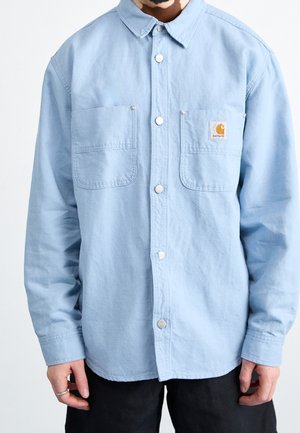 Homme portant une chemise bleu clair à boutons avec deux poches frontales, dont une ornée d'un petit écusson marron avec le logo Carhartt, associée à un pantalon noir.