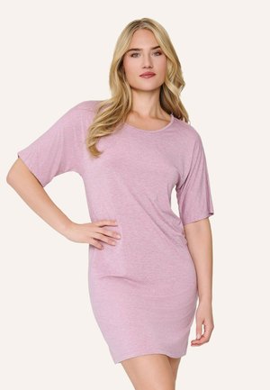 Žena s dlhými blond vlasmi, oblečená v svetlo ružových krátkych rukávoch a neformálnych šatách, pózuje s jednou rukou na bok proti jednoliatemu pozadiu.