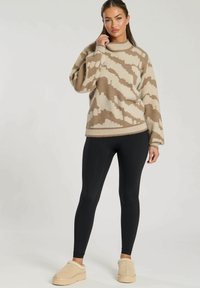 Beżowy i brązowy sweter dzianinowy z wzorem, z wysokim okrągłym dekoltem, w zestawie z czarnymi legginsami i beżowymi butami wsuwanymi. Sweter ma luźny krój.