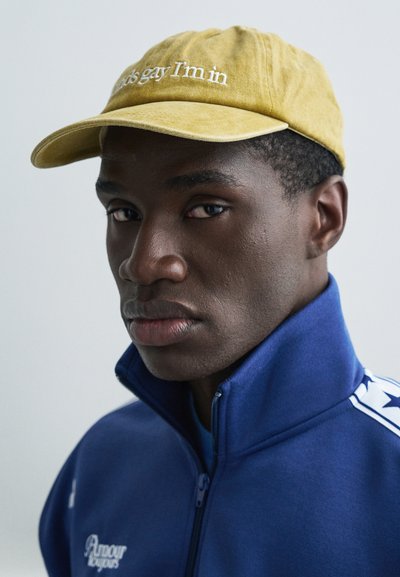 Dzeltens cepure ar baltu tekstu "Es esmu gejs, es esmu iekšā." Zils džemperis ar rāvējslēdzēju un paceltu apkakli, baltas akcenta detaļas. Gluda auduma tekstūra.