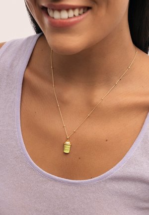 Collar de cadena de oro que presenta un pequeño colgante verde en forma de spray con el texto "ATCHA ERA". El fondo tiene un suave tono lavanda.