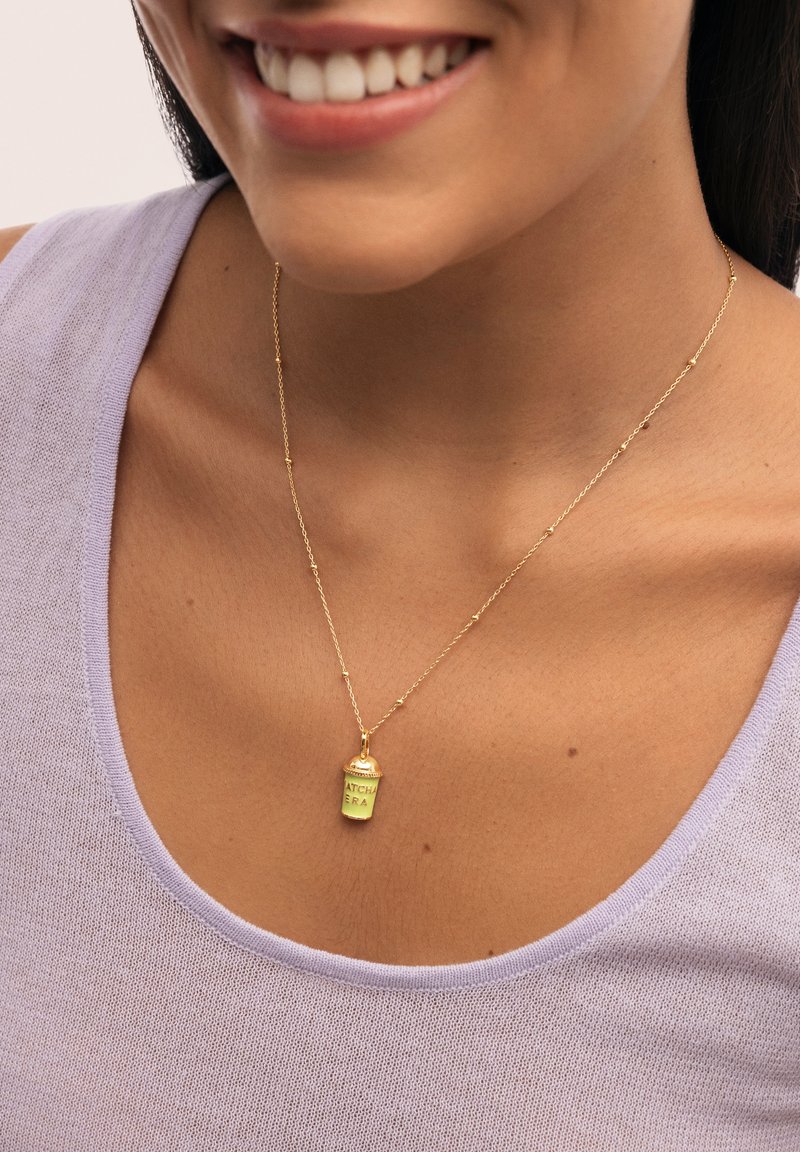 Collar de cadena de oro que presenta un pequeño colgante verde en forma de spray con el texto "ATCHA ERA". El fondo tiene un suave tono lavanda.