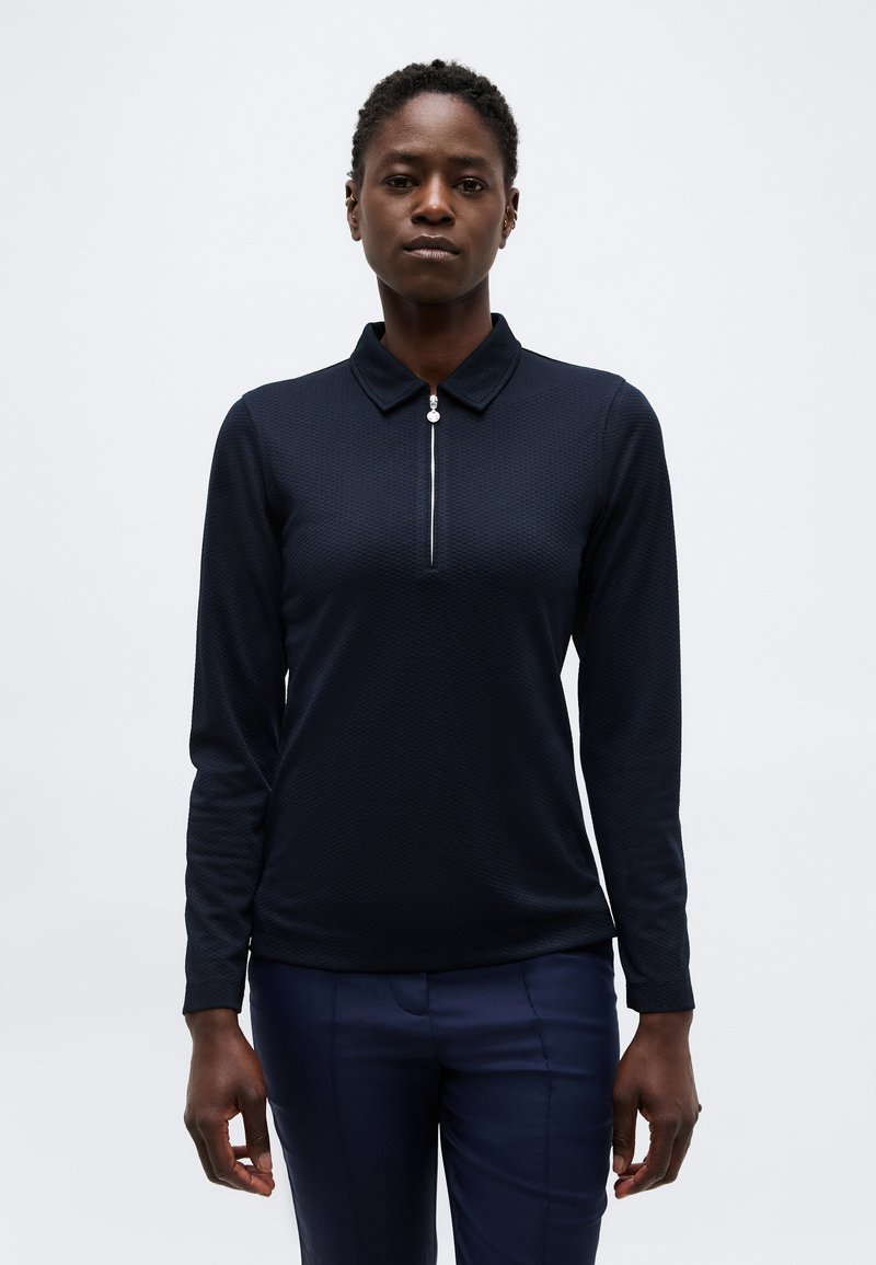 Γυναίκα με κοντά σγουρά μαλλιά φοράει μπλε σκούρο πουκάμισο με μακριά μανίκια, γιακά και φερμουάρ μπροστά, συνδυασμένο με μπλε σκούρο παντελόνι, στέκεται μπροστά σε απλό φόντο.